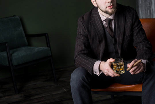 Man in expensive suit is sitting on the leather armchair and drinking whiskey. No face