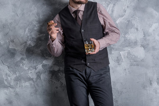 Man in vest standing, leaning against the wall, holding a glass of whiskey. No face
