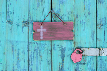 bBlank pink wood sign with a religious cross next to heart lock hanging on rustic antique teal blue wooden door; Easter and Memorial Day holiday concept background with copy space
