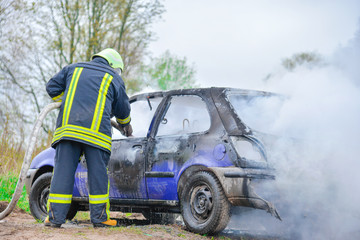 Obraz premium Fire fighter prepare to attack a propane fire. Burning and crashed car after explosion. Accident on street at countryside. No one was injured.