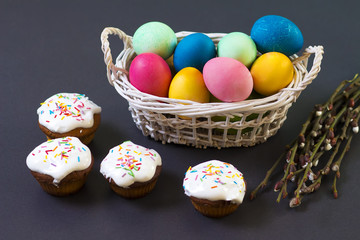 Easter multicolored eggs in a basket and sweet cupcakes muffins on black background. Easter conceptual background for religious Christian holiday.