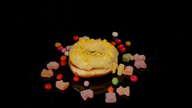 yellow funny surprised glazed donut with sprinkles, striped caramel candies, colorful dragee with raisins or peanuts inside rotate on black background