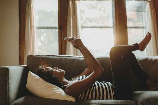 Side View Of Woman Using Smart Phone While Lying On Sofa Against Window At Home