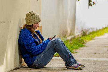 Outdoor view of homeless woman begging on the street in cold autumn weather sitting on the floor at sidewalk with a cellphone in her hand