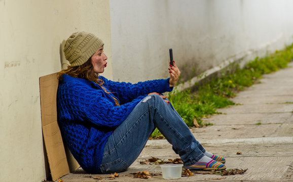 Outdoor view of homeless woman begging on the street in cold autumn weather sitting on the floor at sidewalk with a cellphone in her hand