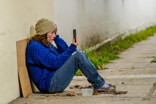 Outdoor view of homeless woman begging on the street in cold autumn weather sitting on the floor at sidewalk with a cellphone in her hand