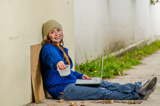 Outdoor view of homeless smiling woman begging on the street in cold autumn weather sitting on the floor with a empty plastic flask and computer over her legs, at sidewalk