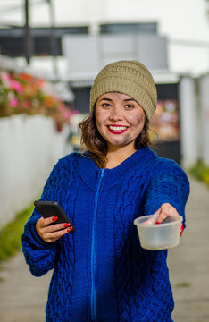 Outdoor view of homeless sad woman on the street in cold autumn weather holding an empty plastic flask in her hands asking for money, using a cellphone at sidewalk