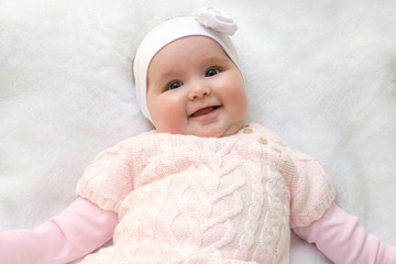 Fototapeta na wymiar Cute cheerful newborn dressed in pink sweater on white fur blanket. Adorable infant baby with headband.