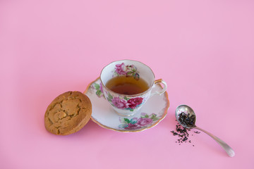 Tea in an antique tea cup with loose leaf tea and a cookie on a pink background