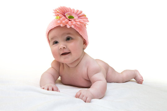 Funny female newborn with nice pink crocheted cap on white background. Happy baby girl with nice knitted pinky cap on white fur blanket. Newborn wearing cute knitted cap with big flower.