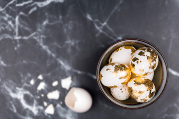 Stylish background, white eggs decorated with golden foil in rustic bowl, and broken egg shell. Space for text on black marble background. Feminine flat lay, narrow depth of field. Easter concept.