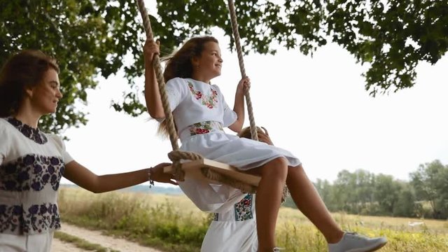 Lovely family. Mother, elder daughter in an embroidered dresses sway a sweet little girl on a wooden swing seat. Family connections, family love. Slow motion