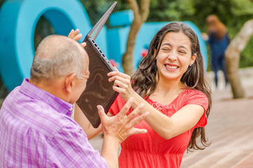 Outdoor view of father and daughter in the park, daugher is going to hit his dad with computer