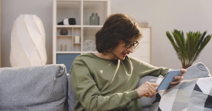 Young handsome man with a long hair scrolling and taping on the tablet device, watching something and laughing on the sofa at home. Indoor