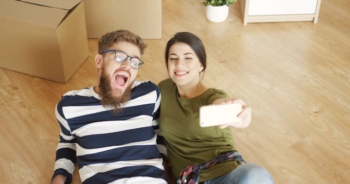 Young joyful couple lying on the floor near boxes and making funny selfies while they moving in the new apartment. Inside