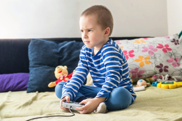 Cute boy playing video game console while sitting on sofa at home. Feeling angry