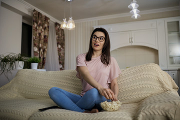 A brunette girl wearing glasses with popcorn watching TV sitting on the sofa in the room.