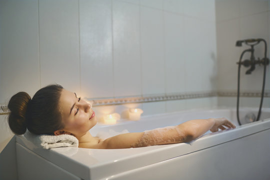 Young girl is resting in a bathtub with foam in bethroom with candles.