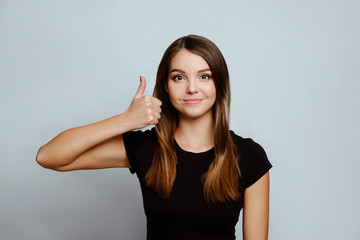 Positive emotions and feelings on white background.Caucasian female with long hair, wearing black T-shirt, showing thumbs up near face and smiling broadly while looking at camera