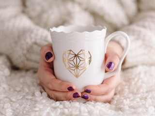 Taza de café con corazón dorado sujetada por manos con uñas moradas
