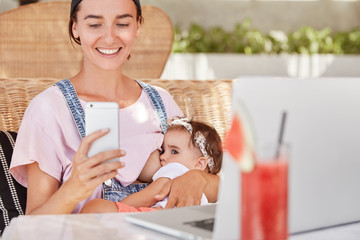 Pretty female, young mother dressed in denim overalls, breastfeeds her newborn child, embraces little daughter, holds smart phone and reads mother`s blog in internet, uses electronic devices