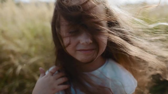 Close up view of cute little girl smiling straight to camera, standing on a strong wind. Summer breeze playing with her hair. Stormy weather, natural beauty. Calmness, relaxation Slow motion