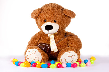 großer brauner Plüsch-Teddy sitzt auf weißem Untergrund und vor weißem Hintergrund umgeben von bunten Ostereiern aus Plastik