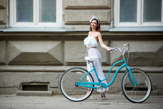pretty young female in long white dress posing with vintage blue bicycle with old historical building on the background. Summer day beautiful girl turquoise bike smiling happy enjoying