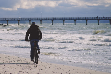 Fototapeta na wymiar A lone cyclist in dark clothes rides on a sandy beach towards the pier.