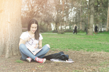 Beautiful young school or college girl with long hair and eye glasses sitting on the ground in the park reading the books and study for exam