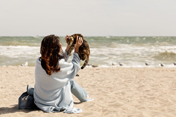 Young woman (brunette) in a light blue cardigan and jeans, with a backpack, sits on the beach and plays with a cat.