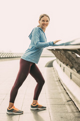 Portrait of smiling young beautiful woman looking at camera, wearing sportswear, leaning on railing and stretching calf on bridge outdoors. Side view. © Mangostar