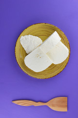 Cheese, Greek cheese on a purple background, feta on a yellow plate, dairy product in minimalism style, copy space, top view, milk breakfast, wooden cutlery, ultraviolet