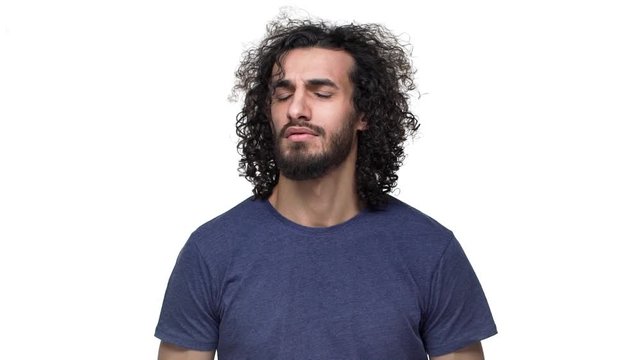 Portrait of hansdome man in casual dark blue t-shirt posing on camera with smile and blowing locks of his dark hair, over white background in slow motion. Concept of emotions