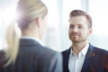 Young confident manager looking at colleague while listening to her speech or opinion at meeting