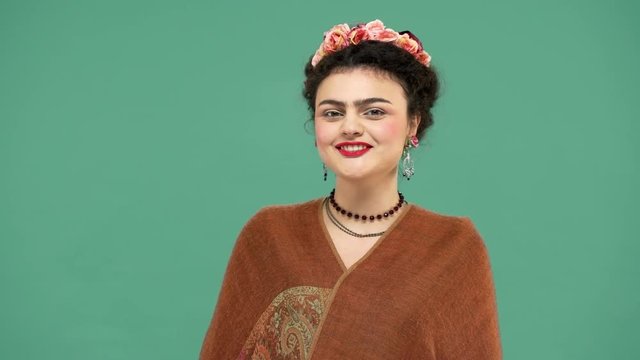 Beautiful portrait of woman with thick eyebrows Frida Kahlo lookalike wearing jewelry and roses in hair smiling with candid artistic, over green background. Stylization concept