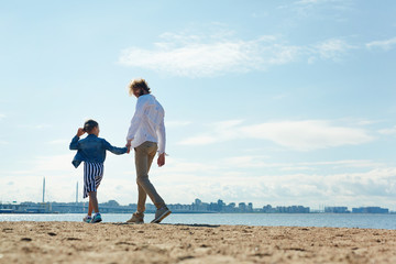 Young man with his daughter walking down sandy beach on warm summer day