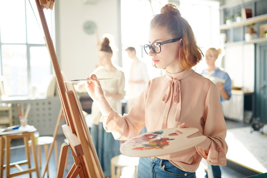 Pretty young woman with palette and paintbrush standing in front of easel and painting
