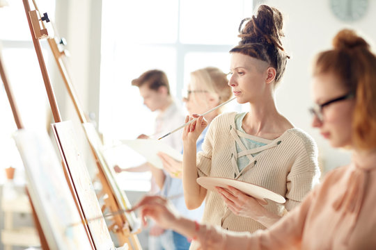 Pensive student with palette and paintbrush looking attentively at painting on easel