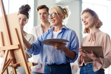 Mature teacher surrounded by group of students showing them painting technique on easel