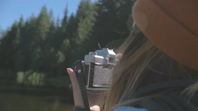 Close up over the shoulder shot of a young woman taking a picture of a mountain with her vintage camera by the lake shore on a sunny day
