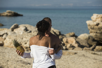 Couple stand at rocky sea beach and kissing on sunny summer day. Couple on vacation hold pineapple. Sexy lady with nude shoulders hugs macho, sea on background. Tropical honeymoon concept. - Powered by Adobe