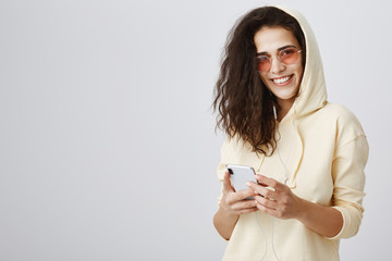 Getting ready to go for walk with music in ears. Portrait of emotive happy young woman in hood and stylish sunglasses listening songs in earphones, holding smartphone and gazing at camera