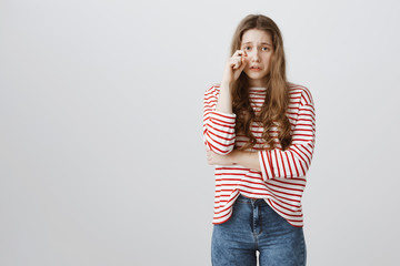 Girl is crying after scolding. Portrait of upset frowning pretty teenager removing tear with hand and whining, being sad and gloomy, partly crossing chest with hand, standing against gray background