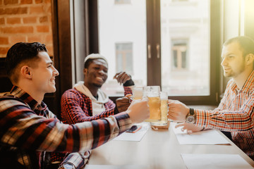 Handsome multiethnic men are clinking cup of beer talking smiling while resting in pub