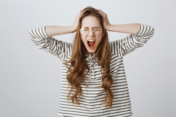 Girl suffers from depression, being under pressure from deadlines and work. Mad cute woman in glasses, screaming or shouting with hands on head, having problems, freaking out over gray wall