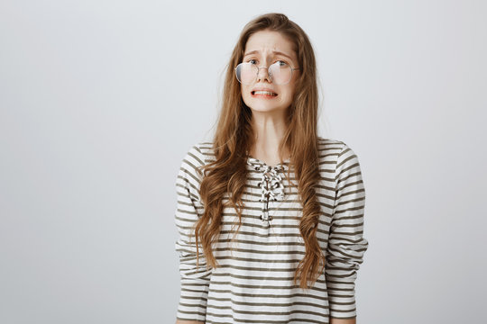 Yikes, bad thing happened. Portrait of confused nervous caucasian teenager in glasses, frowning, clenching teeth and grimacing, feeling embarrassed and awkward, being ashamed over gray wall