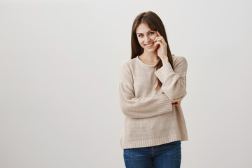 Woman in love is shy to look right in eyes. Portrait of kind good-looking female smiling broadly, standing with partly crossed hands and fingers on face, blushing or enjoying conversation