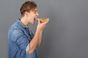 Young man in jeans jacket studio isolated on grey holding pizza sice side view